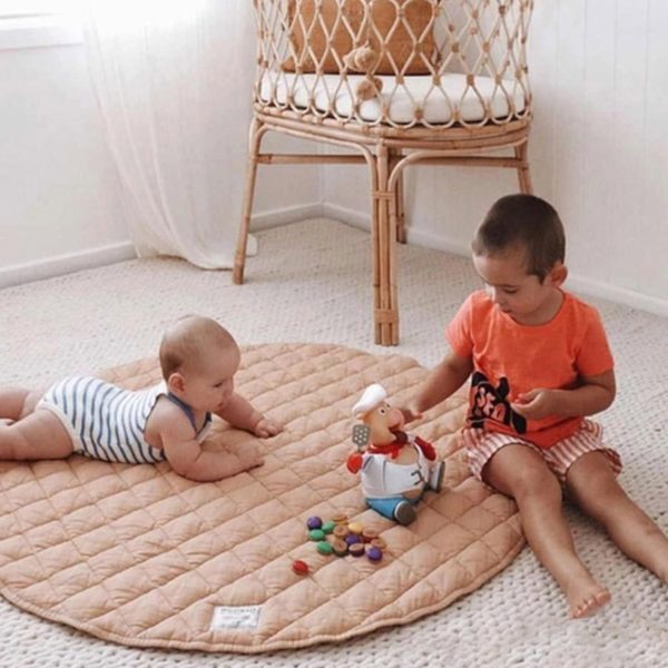 tapis de sol rond rose , bébé et enfant qui jouent dessus et une chaise en rotin avec un coussin blanc