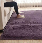 Tapis doux rectangulaire violet installé au pied d'un canapaté, on voit des jambes avec des chaussons de la personne assise sur le canapé