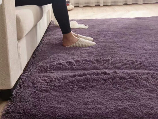 Tapis doux rectangulaire violet installé au pied d'un canapaté, on voit des jambes avec des chaussons de la personne assise sur le canapé